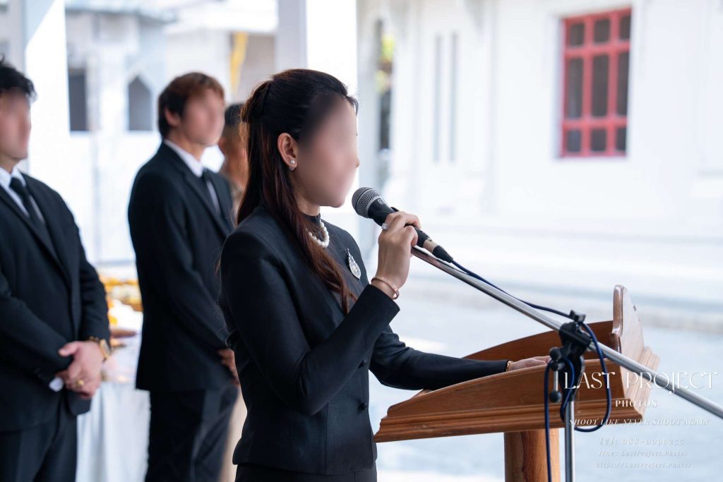 ภาพถ่ายระยะใกล้เจ้าภาพหญิงกำลังกล่าวคำไว้อาลัยแด่ผู้วายชนม์ด้วยความสงบสำรวมในพิธีฌาปนกิจ Close-up shot of a female host respectfully reading a eulogy at the podium during a Thai cremation ceremony