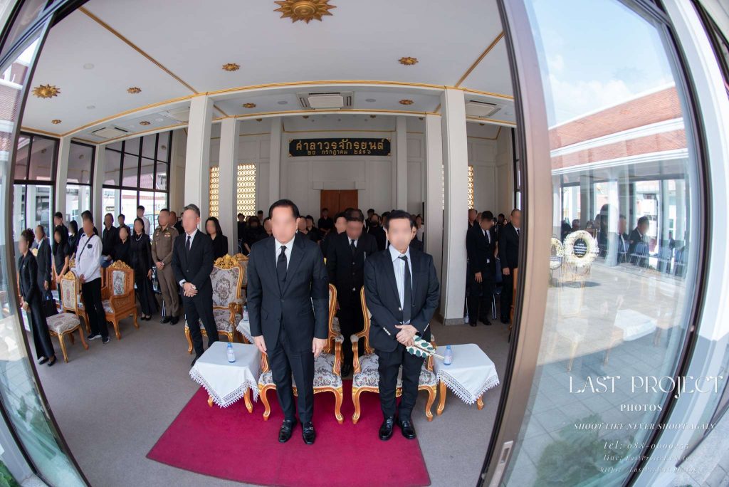 แขกผู้มีเกียรติยืนสงบนิ่งไว้อาลัยในพิธีฌาปนกิจศพ ณ ศาลาวัด ถ่ายภาพมุมกว้างแบบสมมาตร Guests standing in silent tribute during a Buddhist cremation ceremony at a Thai temple pavilion captured with a wide-angle lens
