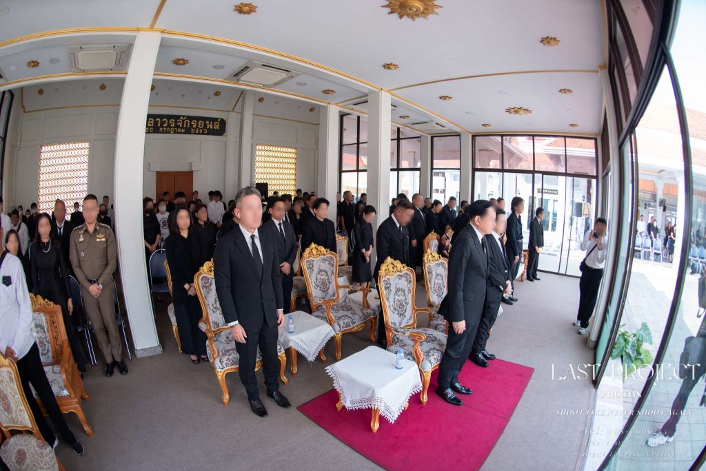 แขกผู้มีเกียรติและญาติยืนสงบนิ่งร่วมไว้อาลัยในพิธีฌาปนกิจศพ ณ ศาลาวัด ถ่ายด้วยเลนส์มุมกว้างพิเศษ Guests and mourners observing a solemn moment of silence during a Buddhist cremation ceremony inside a Thai temple pavilion captured with an ultra-wide lens