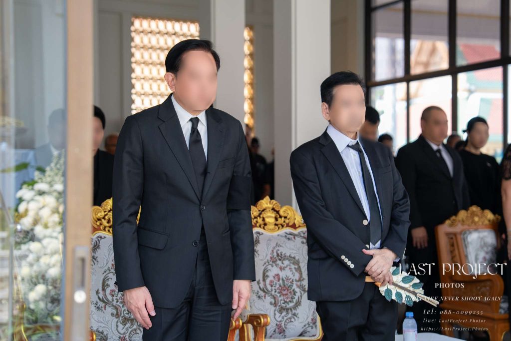 แขกผู้มีเกียรติในชุดสูทสีดำร่วมไว้อาลัยในพิธีฌาปนกิจศพ ณ วัดไทย Guests in black formal suits standing respectfully during a Thai Buddhist cremation ceremony at a temple