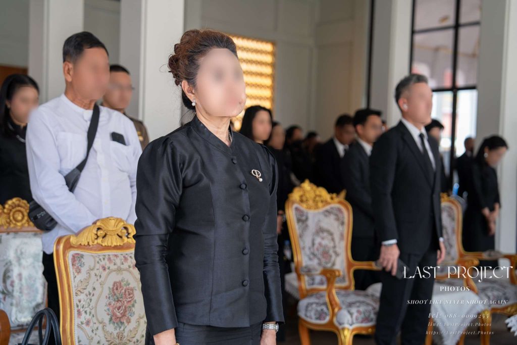 แขกผู้มีเกียรติสวมชุดไว้ทุกข์สีดำยืนสงบนิ่งไว้อาลัยในพิธีฌาปนกิจศพ ณ วัดไทย Guests in black mourning attire standing in a moment of silence during a Thai Buddhist cremation ceremony at a temple