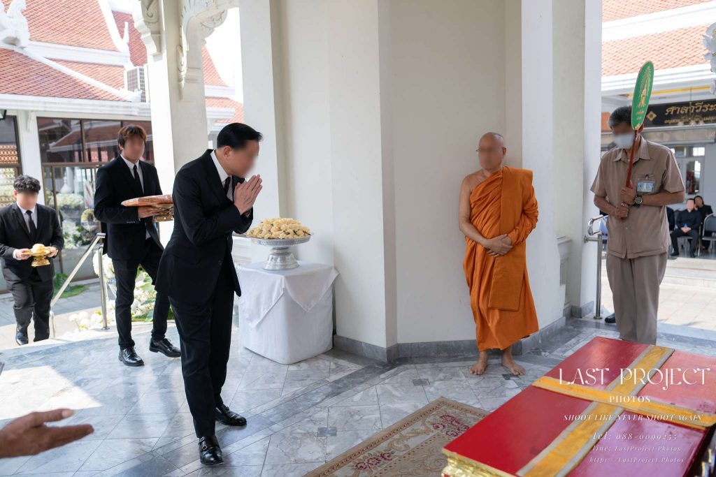 แขกในชุดสูทสีดำยกมือไหว้ทำความเคารพศพและพระสงฆ์บนเมรุในพิธีฌาปนกิจ A guest in a black suit paying respects with a wai to the deceased and a Buddhist monk at the crematorium during a Thai cremation ceremony.