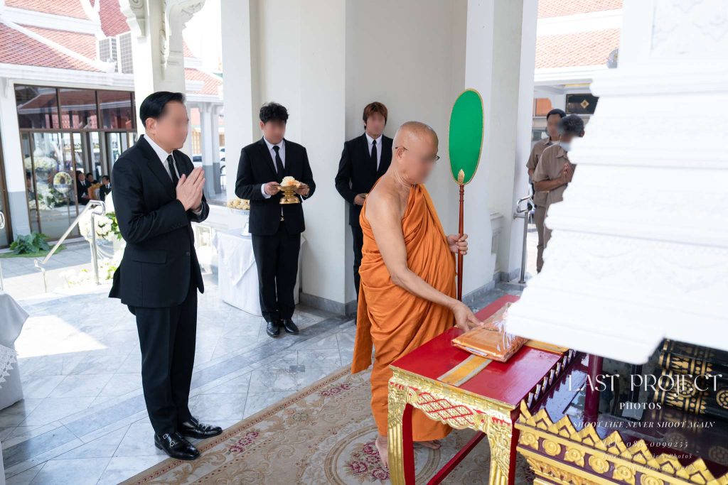 แขกผู้มีเกียรติยืนพนมมือขณะพระสงฆ์พิจารณาผ้าไตรบังสุกุลบนเมรุในพิธีฌาปนกิจศพ ณ วัดไทย Distinguished guest with hands in prayer while a senior Buddhist monk contemplates the final Bangsukula robe on the crematorium platform during a Thai cremation ceremony at a temple
