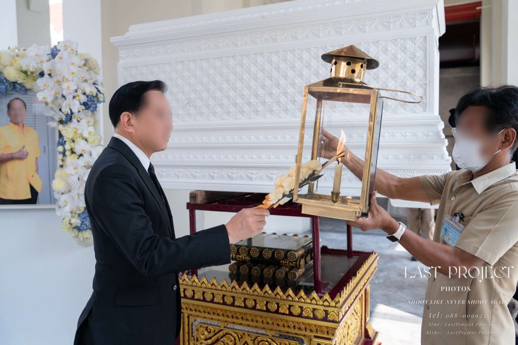 เจ้าภาพจุดดอกไม้จันทน์จากตะเกียงไฟในพิธีฌาปนกิจศพ โดยมีภาพถ่ายผู้วายชนม์เป็นฉากหลัง Host lighting a sandalwood flower from a flame lantern during a Thai Buddhist cremation ceremony with the deceased portrait in the background