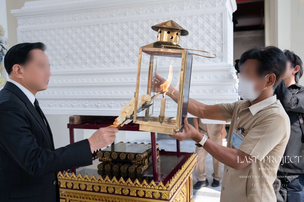 ภาพประธานในพิธีจุดไฟดอกไม้จันทน์จากตะเกียงหน้าหีบศพ ในพิธีฌาปนกิจศพ ณ วัดไทย A guest lighting a sandalwood flower from a ceremonial flame lantern before the coffin during a Thai Buddhist cremation ceremony.