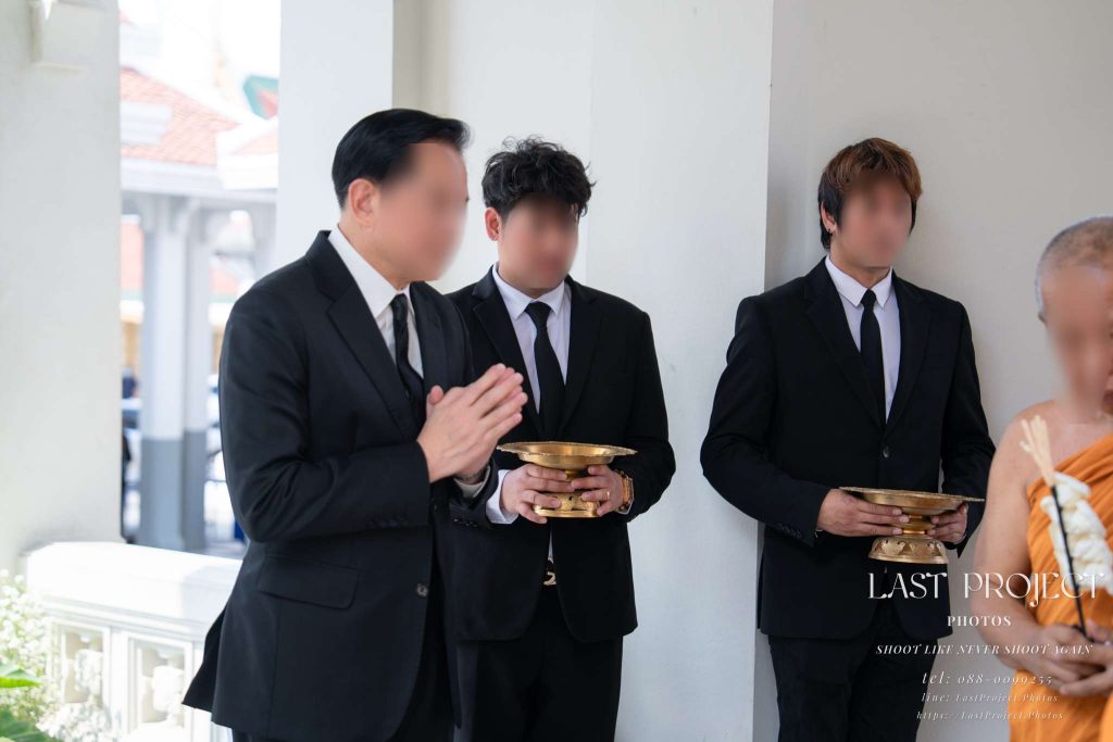 เจ้าภาพยืนพนมมือแสดงความเคารพระหว่างพระสงฆ์วางดอกไม้จันทน์ในพิธีฌาปนกิจ Hosts with hands clasped in prayer paying respects as monks place sandalwood flowers at a Thai cremation ceremony