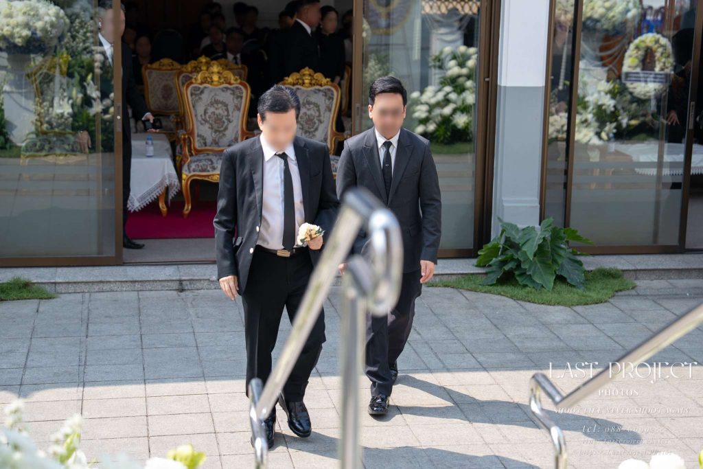 แขกสวมชุดสูทสีดำถือดอกไม้จันทน์เดินลงบันไดในพิธีฌาปนกิจศพที่วัดไทย Guests in black suits holding traditional sandalwood flowers walking down the steps during a Thai Buddhist cremation ceremony at a temple