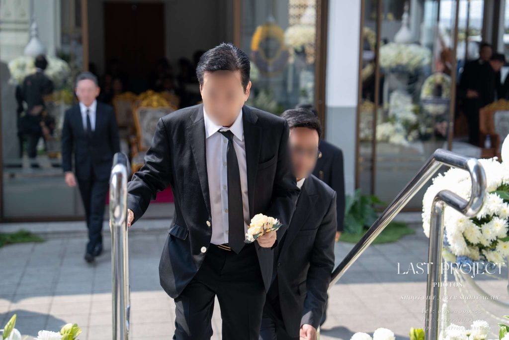 ภาพถ่ายเจาะแขกผู้มาร่วมงานถือดอกไม้จันทน์เดินขึ้นบันไดเมรุในพิธีฌาปนกิจศพที่วัดไทย Close-up shot of a funeral guest holding a sandalwood flower while walking up the crematorium stairs at a Thai Buddhist cremation ceremony