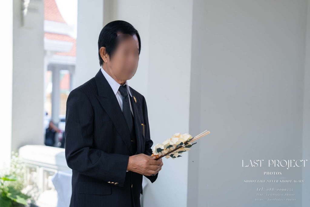 แขกอาวุโสในชุดสูทสีดำถือดอกไม้จันทน์ด้วยความสำรวมบนเมรุในพิธีฌาปนกิจศพ Elderly guest in a black suit reverently holding sandalwood flowers on the crematorium terrace during a Thai Buddhist cremation ceremony